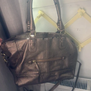 Bronsfärgad handväska med axelrem - Säljer en bronsfärgad handväska med både handtag och avtagbar axelrem. Väskan har en dragkedja framtill och detaljer i guld. Tillverkad i syntetmaterial med struktur och flera fack för förvaring. Perfekt för dig som vill ha en rymlig och snygg accessoar. (sliten, se på bilderna) 