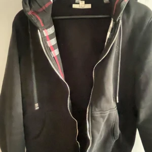 Svart hoodie med rutigt foder i huvan - Säljer en svart hoodie från Burberry med dragkedja och klassiskt rutigt foder i huvan. Tröjan har fickor framtill och justerbara snören i huvan. Perfekt för en stilren och avslappnad look.