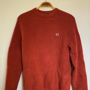 Rostfärgad stickad tröja från Fred Perry - Säljer en snygg rostfärgad stickad tröja från Fred Perry med rund halsringning och diskret logga på bröstet. Tröjan har lång ärm och ribbade muddar vid ärmslut och nederkant. Perfekt för kyligare dagar!