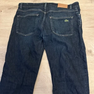 Mörkblå jeans från Lacoste - Säljer ett par mörkblå jeans från Lacoste med klassisk femficksdesign och dragkedja. Byxorna har raka ben och Lacostes ikoniska krokodillogga på bakfickan. Perfekta för dig som gillar stilrena och tidlösa jeans. Storlek -32/34, skick- nyskick.