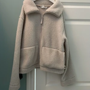 Beige teddyjacka från H&M - Mysig beige teddyjacka från H&M med dragkedja framtill och två stora fickor. Jackan har lång ärm och hög krage, perfekt för kyliga dagar. Passar dig som gillar en avslappnad och trendig stil. Andrig använd