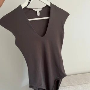 Mörkgrå/brun v-ringad bodysuit - Säljer en stilren mörkgrå/brun bodysuit med v-ringning och ärmlös design. Mjukt och stretchigt material för skön passform.