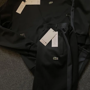 Svart träningsjacka från Lacoste - Säljer en svart träningsjacka från Lacoste med dragkedja framtill och ribbade muddar. Jackan har diskret Lacoste-logga på bröstet och sportiga detaljer längs ärmarna. Perfekt för dig som gillar stilren och sportig look.