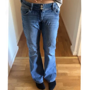 Blå bootcut jeans med låg midja - Säljer ett par klassiska blå jeans med bootcut-modell och låg midja, dubbla knappar och dragkedja framtill. Perfekta för en avslappnad och tidlös look.