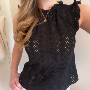 Svart ärmlös blus med spetsdetaljer - Svart blus med broderade blommor och spetsdetaljer. Ärmlös modell med volang vid axlarna och hög halsringning. Perfekt för dig som gillar romantisk och feminin stil.