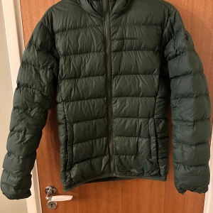Grön dunjacka från Peak Performance - Mörkgrön dunjacka från Peak Performance med huva och dragkedja framtill. Jackan har quiltad design och är perfekt för kyliga dagar. Klassisk passform och diskret logga på bröstet och ärmen.