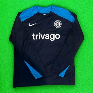 Chelsea långärmad fotbollströja från Nike - Chelsea långärmad träningströja från Nike med blå och svarta paneler. Tröjan har klubbmärke på bröstet, vit Nike-logga och sponsortryck 'trivago' på framsidan. Perfekt för fotbollsträning eller support.