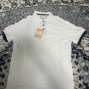 Vit pikétröja från Johnson & Sons - Säljer en klassisk vit pikétröja från Johnson & Sons i storlek XL. Tröjan har svarta detaljer vid ärmslut och krage samt knappar framtill. Perfekt för en stilren look och tillverkad i bomull för extra komfort.