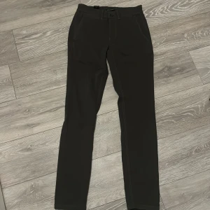 Mörkbruna chinos från Lindbergh - Säljer ett par mörkbruna chinos från Lindbergh med klassisk passform. Byxorna har sidfickor, bälteshällor och knäppning fram. Materialet är bomull och de har diskreta bakfickor med knappar. Perfekta för en stilren look. Ny pris på dess byxor går för 800kr