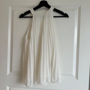Vit plisserad topp - Säljer en vit plisserad topp med ärmlös design och rund halsringning. Toppen har en luftig och flowy passform som ger en elegant känsla. Perfekt för sommarens tillfällen och midsommarfirande.
