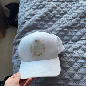 Vit keps från BOSS - Snygg vit keps från BOSS med guldigt tryck på framsidan där det står 'BOSS' i en cirkel. Klassisk böjd skärm och justerbar passform baktill. Perfekt accessoar för en stilren look.