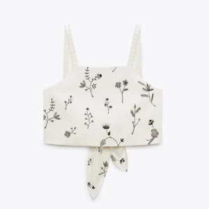 Vit blommig linnetopp med brodyr knyt - Superfin vit linnetopp med tunna axelband och svart blommigt mönster. Toppen har en snygg knytning framtill som ger en extra detalj