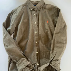 Olivgrön skjorta från Ralph Lauren - Klassisk olivgrön skjorta från Ralph Lauren med knappar framtill och broderad logga på bröstet. Skjortan har lång ärm och button-down krage, perfekt för en stilren look.