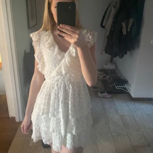 Vit spetsklänning med volanger - Superfin vit kortklänning i spets med volangdetaljer och omlottliv från Nelly. Klänningen har v-ringning och är ärmlös med romantiska volanger på axlarna. Perfekt för sommarens alla tillfällen! Klänningen är helt oanvänd. 