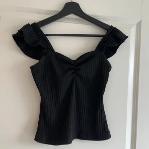 Svart ribbad topp med volangärmar - Säljer en svart ribbad topp med breda axelband och volangdetaljer vid axlarna. Toppen har en lätt v-ringning och smickrande passform. Perfekt att styla till olika outfits!