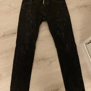 Svarta jeansbyxor från Dsquared2 - Svarta jeansbyxor från Dsquared2 med diskreta slitningar och klassisk femficksdesign. Byxorna har dragkedja och knapp framtill samt Dsquared2-logga på insidan. Snygg, smal passform som passar till många stilar. Men vänstra fickan har ett litet hål och fickan åt höger har också ett litet större hål men du kan fortfarande förvara saker där som mobil nycklar etc