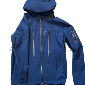 Blå vindjacka  - Snygg blå vindjacka från revolution race med huva och flera dragkedjeförsedda fickor. Jackan har en sportig look och är perfekt för utomhusaktiviteter. Justerbar huva och ärmslut för extra komfort.