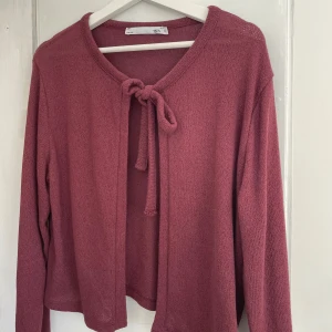 Rosa kofta med knyt från 157 - Säljer en söt rosa kofta från 157 i storlek M. Koftan är stickad och har långa ärmar samt ett knyt framtill vid halsen. Perfekt att slänga över en topp för en avslappnad look.