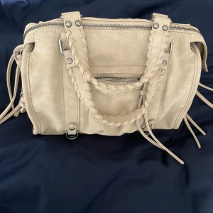 Beige axelväska med flätade handtag - Säljer en beige axelväska med snygga flätade handtag och silverfärgade detaljer. Väskan har dragkedja upptill, flera fack och en avtagbar axelrem. 