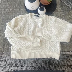 Vit stickad tröja med kabelmönster - Säljer en vit stickad tröja med snyggt kabelstickat mönster och rund hals. Tröjan har långa ärmar och breda ribbade muddar vid ärmslut och nederkant. Perfekt för kyliga dagar nu i sommar!