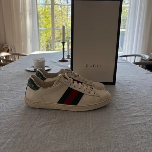 Gucci ace - Säljer mina Gucci Ace sneakers i storlek 42. Skorna är i vitt läder med det ikoniska gröna och röda sidobandet, samt grön metallic-detalj baktill. De är sparsamt använda och i mycket fint skick, allt og (originalkartong, dust bag, extra sko snören och kvitto) medföljer. 	•	Storlek: 42 	•	Färg: Vit med grönt/rött band 	•	Skick: Mycket bra skick, se bilder 	•	Nypris: ca 6.500 kr  Perfekta sneakers för dig som vill kombinera lyx och stil både till vardags och fest.  Hör av dig vid minsta fundering😊