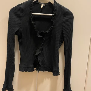 Svart ribbad tröja från NLY Trend - Säljer en svart ribbad kofta från NLY Trend i storlek M. Koftan har långa ärmar, volangkant längs öppningen, ärmslut och nederkant. 