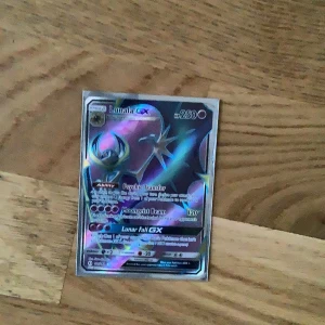 Pokémonkort Lunala GX - Säljer ett holografiskt Pokémonkort av Lunala GX med 250 HP. Kortet har färger i lila, blått och silver med glansig yta och detaljerad illustration. Perfekt för samlare eller Pokémonfans som vill utöka sin samling.