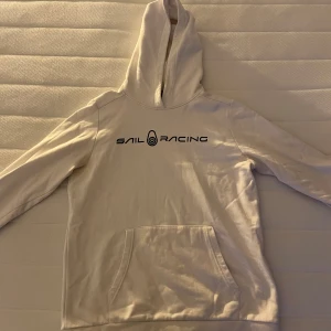 Vit hoodie från Sail Racing - Vit hoodie från Sail Racing med svart logga på bröstet. Tröjan har huva och en stor magficka framtill. Den är nästan helt ny och är i storlek 176! Ifall det är några frågor så är det bara att skicka!