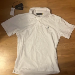 Vit piké från Polo Ralph Lauren - Klassisk vit pikétröja från Polo Ralph Lauren med broderad logga på bröstet. Tröjan har krage och knappar framtill, perfekt för en clean och stilren look. Tillverkad i mjuk bomull för extra komfort. 