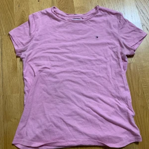 Rosa t-shirt från Tommy Hilfiger - Säljer en klassisk rosa t-shirt från Tommy Hilfiger med liten broderad logga på bröstet. T-shirten har rund halsringning och korta ärmar. Perfekt basic-plagg till garderoben!