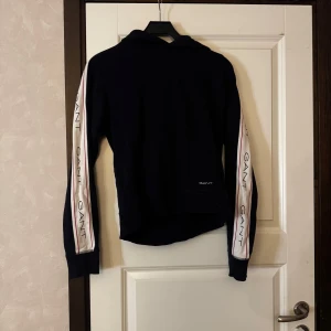 Mörkblå hoodie från GANT med logga på ärmarna - Säljer en mörkblå hoodie från GANT med vita och röda ränder längs ärmarna där GANT-loggan är tryckt flera gånger. Tröjan har huva och långa ärmar, perfekt för en avslappnad stil.