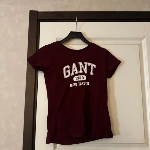 Vinröd t-shirt från GANT - Säljer en vinröd t-shirt från GANT med vit tryckt text 'GANT 1949 NEW HAVEN' på bröstet. Klassisk rund halsringning och korta ärmar. Perfekt för en avslappnad stil.