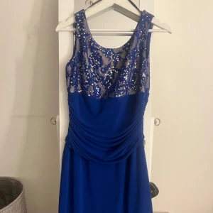 Blå långklänning med paljetter - Säljer en elegant blå långklänning med glittriga paljetter upptill och draperad midja. Klänningen är ärmlös och har en rund halsringning. Perfekt för bal eller festliga tillfällen.