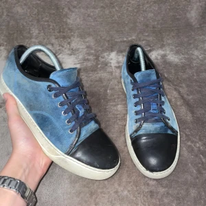 Lanvins  - Säljer ett par blå sneakers med svart tå och svarta detaljer. Skorna har snörning och vit sula. Ovandelen är i mocka och tån är i skinn. Perfekta för dig som gillar en stilren och avslappnad look.