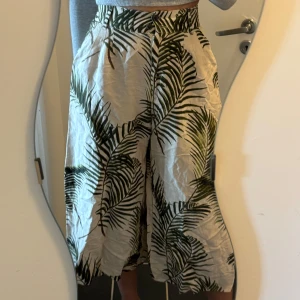 Beige trekvarts byxor med palmbladsmönster - Säljer en beige trekvarts byxa med grönt palmbladsmönster. Byxorna har hög midja och en luftig, vid passform som ger en skön sommarkänsla. Är 177 och dom slutar ungefär vid vaden.