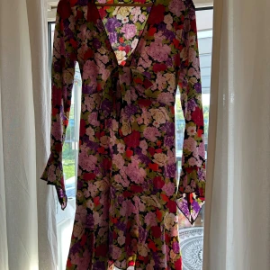 Blommig långärmad klänning med knyt från Gina Tricot - Färgglad blommig klänning från Gina Tricot med v-ringning och knyt framtill. Långärmad modell med volangdetaljer nertill och i ärmslut. Mönstret har lila, rosa, rött, grönt och vitt.