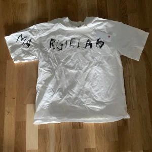 Vit t-shirt från Maison Margiela MM6 - Vit t-shirt från Maison Margiela MM6 med svart handmålat tryck på bröstet och ärmen. Oversized passform och rund halsringning. Tillverkad i Italien, perfekt för en trendig och avslappnad stil.