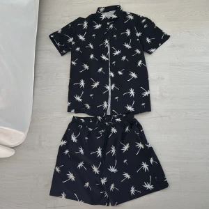 Svarta shorts och svart skjorta med palmtryck - Svarta shorts och svart skjorta med vita palmträdsmönster och elastisk midja med snörning. Perfekta för sommaren och har en avslappnad passform. Materialet känns lätt och svalt, perfekt för varma dagar.   !! Shortsen kan köpas separat !!