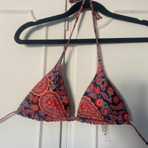 Färgglad mönstrad trekantsbikini - Säljer en trekantsbikini med färgglatt blommigt mönster i rött, blått och orange. Bikinin har knyt i nacken och ryggen samt volangkant nedtill. Perfekt för sommardagar på stranden!