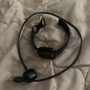 Svart smartklocka från Fitbit med laddare - Säljer en svart smartklocka från Fitbit med tillhörande USB-laddare. Klockan har en fyrkantig display och silikonarmband med flera justeringshål. Perfekt för dig som vill hålla koll på hälsa och aktivitet.