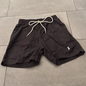 Svarta shorts från Polo Ralph Lauren - Svarta shorts från Polo Ralph Lauren med vit broderad logga framtill. De har elastisk midja med vit snörning och två bakfickor. Perfekta för sommaren och har en klassisk, enkel stil.