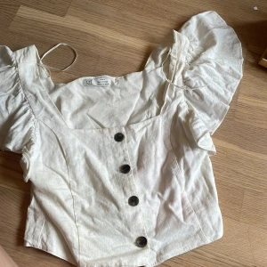 Vit croppad blus med volangärm från Zara - Säljer en vit croppad blus från Zara med stora volangärmar och knappar framtill. Blusen har en fyrkantig urringning och är tillverkad i ett lätt linnematerial. Perfekt för varma dagar och ger en trendig look.