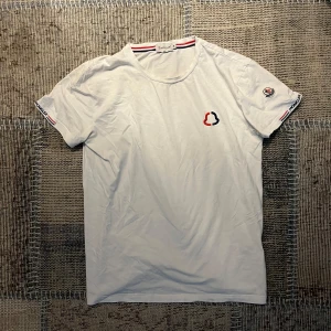 Vit t-shirt från Moncler - Säljer en vit t-shirt från Moncler med broderad logga på bröstet och ärmen. T-shirten har korta ärmar och snygga röd-blå-vita detaljer vid halsen och ärmslut. Perfekt för dig som gillar stilrena märkesplagg.