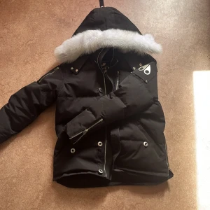 Svart dunjacka med pälskant från Moose Knuckles NY - Säljer en svart dunjacka från Moose Knuckles med stor huva och vit pälskant. Jackan har dragkedja, knappar och flera fickor, samt detaljer i metall. Perfekt för kalla dagar och riktigt snygg streetstil.