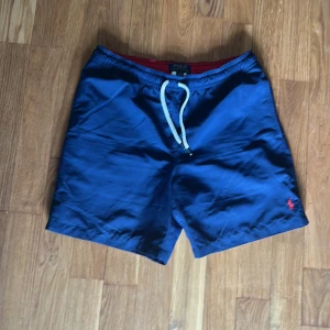 Blå badshorts från Polo Ralph Lauren - Snygga blå badshorts från Polo Ralph Lauren med vit snörning i midjan och röd logga broderad på benet. Shortsen har elastisk midja och en bakficka med knapp. Perfekta för stranden eller poolen.