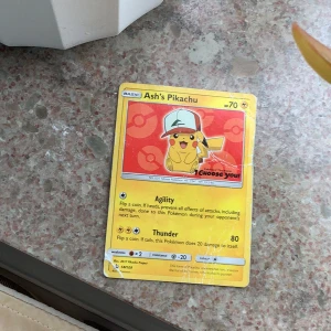 Pokémonkort Ash's Pikachu - Säljer ett samlarkort från Pokémon med motivet Ash's Pikachu. Kortet har en gul ram, Pikachu med Ashs keps och texten 'I Choose You!'. Kortet har 70 HP och attacker som Agility och Thunder. Perfekt för samlare eller Pokémon-fans.