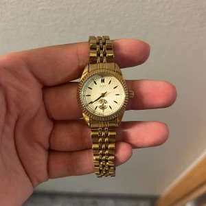 Guldfärgad klocka från Regal - Snygg guldfärgad klocka från Regal med klassisk rund urtavla och länkarmband i metall. Urtavlan har en stilren design med små indexmarkeringar och en extra liten sekundvisare. Perfekt accessoar för dig som gillar en elegant och tidlös look.