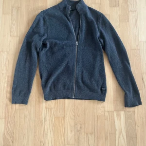 Mörkgrå stickad kofta med dragkedja - Snygg mörkgrå stickad kofta från Jack and Jones med hel dragkedja framtill och ribbade muddar. Perfekt för lager-på-lager och har en stilren look med hög krage. Materialet är mjukt och håller dig varm under kyliga dagar.