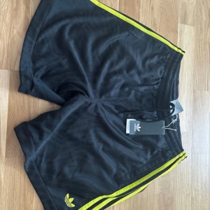 Svarta Adidas shorts med gula ränder - Adidas shorts i svart med subtilt mönster och ikoniska gula ränder längs sidorna. Elastisk midja för skön passform och klassisk Adidas-logga framtill. Perfekta för sport eller chill, tillverkade i lätt polyester som andas. Orginal pris 529!