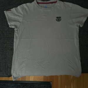 Grå FC Barcelona t-shirt i bomull - Snygg grå t-shirt från FC Barcelona med klubbens emblem tryckt på bröstet. Klassisk rund hals och korta ärmar, med en diskret röd detalj på insidan av kragen. Perfekt för dig som gillar fotboll och vill visa vilket lag du supportar.